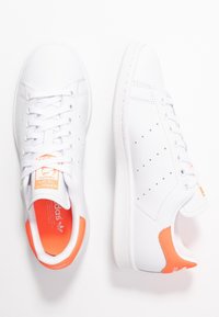 White athletic shoes with a smooth leather upper, perforated detailing, orange accents on the heel and inner lining, and white rubber sole.