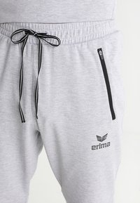 Graue Jogginghose mit schwarzen Kordeln, ausgestattet mit einer seitlichen Reißverschlusstasche und einem schwarzen "erima"-Logo am linken Oberschenkel. Weiche Stofftextur.