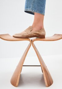 Beige suède instappers met een platte hak, voorzien van een gestikt ontwerp en een licht afgeronde neus, geplaatst op een kromme houten kruk.