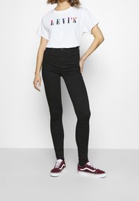Camiseta blanca con logo "LEVI'S" multicolor, pantalones ajustados negros y zapatillas burdeos con detalles en blanco. Pose de pie frente a un fondo liso.