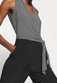 Svart jumpsuit med halterneck, en figurnära topp med rutigt mönster och en knytande midjeaccent. Tillverkad av ett slätt tyg.