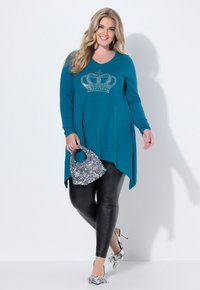 Długi rękaw w kolorze morskim z wzorem korony z kryształków, połączony z błyszczącymi czarnymi legginsami i srebrnymi butami z ostrym noskiem. Mała torebka z cekinami.