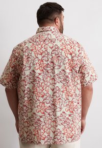 Chemise à manches courtes en tissu léger avec un fond crème, ornée d'un motif floral rouge complexe et d'un détail de col.