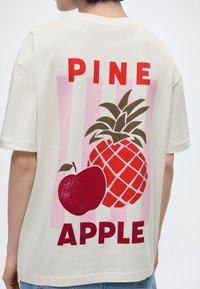 Άτομο που φοράει εκρού μπλουζάκι με κόκκινο κείμενο "PINE APPLE" και μεγάλο γραφικό ενός ανανά και ενός μήλου στην πλάτη.