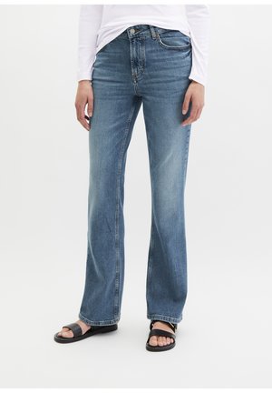 Personne portant un jean en denim bleu taille mi-haute avec une coupe droite aux jambes et des sandales plates noires, debout devant un fond blanc uni.