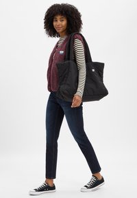 Bolsa tote negra y acolchada, chaleco de forro polar burdeos texturizado sobre una camiseta de manga larga a rayas, jeans oscuros y zapatillas negras con suelas blancas.