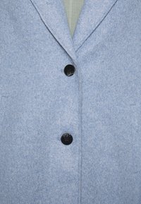 Cappotto in misto lana azzurro chiaro con rever a punte, due bottoni neri e tasche laterali. Tessuto morbido e texture con design minimal.