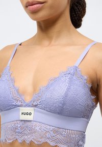 Lavendli pitsist spordirinnahoidja, millel on V-kaelus, lainelised servad ja pehme elastne pael, mille keskel asub "HUGO" silt.