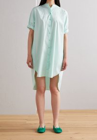 Robe chemise vert clair avec un col mao, des manches courtes retroussées et des fentes latérales, associée à des chaussures plates vertes sur un parquet en bois.