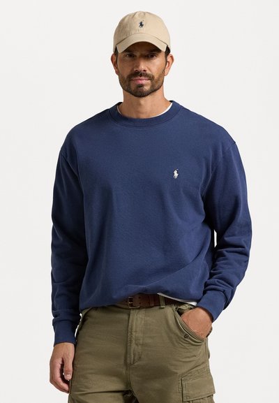 Navy blue cotton sweatshirt dengan lengan panjang dan manset bergaris, menampilkan logo bordir kecil berwarna putih di dada. Dipadukan dengan celana kargo khaki.