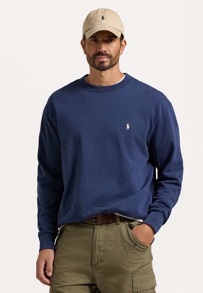 Sweatshirt de algodão azul marinho com mangas longas e punhos canelados, apresentando um pequeno logotipo branco bordado no peito. Usado com calças cargo caqui.