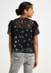 Schwarze, transparente Bluse mit Sterne-Muster, Rüschenärmeln und einer Schlüssel-Aussparung im Rücken, kombiniert mit grauen Jeans, die dekorative Gesäßtaschen haben.