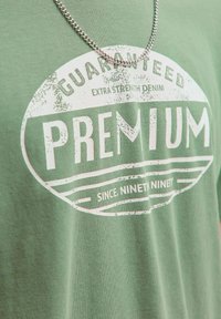 Žalias medvilninis marškinėlis su baltu grafiniu spaudiniu, kuriame užrašyta "PREMIUM", apsuptas apvalaus dizaino ir tekstų "GUARANTEED" bei "SINCE NINET NINETY".