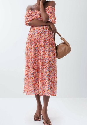 Kvinde i farverig off-shoulder midi-kjole holder en tan håndtaske, iført brune sandaler med hæl på en ensartet lys baggrund.
