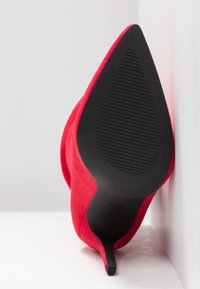 Tacón de aguja de ante rojo con punta afilada, que presenta una suela de goma negra texturizada y un tacón delgado y cónico para una silueta elegante.