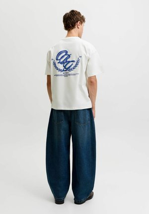 Personne debout de dos portant un t-shirt blanc à motif graphique et un jean large bleu foncé sur fond blanc uni.