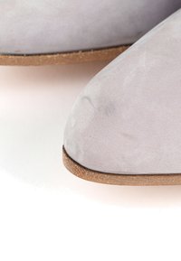 Gros plan sur le bout d'une chaussure en suède gris clair avec une forme arrondie et une fine semelle marron sur fond blanc.