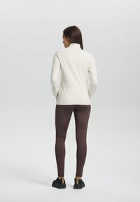 Woman standing with back to camera wearing a white long-sleeve top, brown leggings, and black sneakers on a plain background.