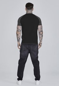 Schwarzes Kurzarm-T-Shirt mit grauen Schulterpaneelen, kombiniert mit dunklen Denim-Jeans. Weiße Sneakers runden das Outfit ab. Strukturierter Stoff sichtbar.