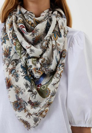 Femme portant un chemisier blanc avec des manches bouffantes et un grand foulard à motif floral noué autour du cou.