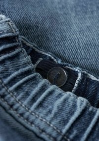 Tessuto di denim di tonalità blu scuro, che mostra un primo piano di un bottone e dettagli di cuciture. La texture appare morbida con una leggera lucentezza.