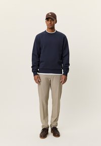 Μπλε σκούρο φούτερ με ριμπ μανσέτες και γιακά, συνδυασμένο με ανοιχτό μπεζ παντελόνι και καφέ παπούτσια. Ένα καφέ καπέλο ολοκληρώνει την εμφάνιση.