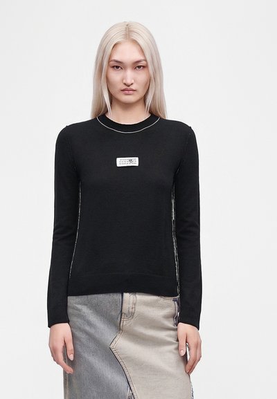 Jeune femme aux longs cheveux blonds portant un pull noir à manches longues et une jupe en jean bicolore, debout devant un fond blanc uni.