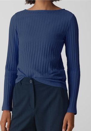 Frau trägt ein blaues, geripptes Langarmshirt, das in hoch taillierte dunkelblaue Hosen mit Knopfverschluss gesteckt ist.