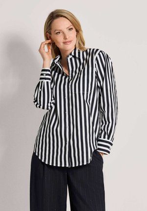 Blonde vrouw met grote creolen en een zwart-wit verticaal gestreept overhemd met knopen en een zwarte broek, poseert zijwaarts.