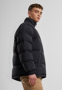 Jachetă puff în negru, cu un guler înalt, textură matlasată și manșete elastice. Asortată cu pantaloni bej care prezintă detalii cu logo.