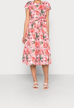 Vrouw in een roze midi-jurk met bloemenprint, kapmouwtjes en een strikceintuur, gecombineerd met witte sandalen met bandjes.