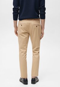 Bež chinos hlače s slim fit krojem, ki imajo dva zadnja žepa s gumbom. Oblikovan z temno morsko modrim puloverjem in črnimi čevlji.