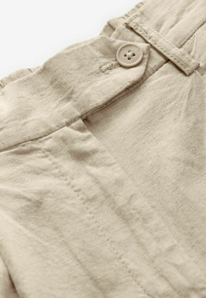 Tessuto di lino beige con chiusura a bottoni visibile e dettagli di cucitura. La texture è leggermente stropicciata, mostrando una finitura morbida e naturale.
