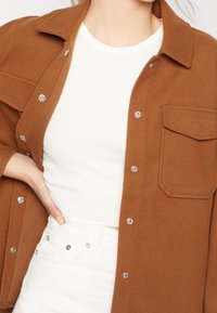 Brun ullblandad skjortjacka med tryckknappar, krage och bröstficka, lager på en vit ribbad linne och vita jeans.