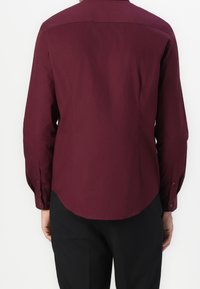 Bordowa koszula z długim rękawem, z wywiniętym kołnierzykiem, zapinanymi mankietami i zaokrąglonym dołem. Tkanina wydaje się gładka o jednolitej teksturze.