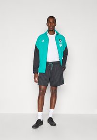 Sportoutfit bestående af en teal og sort zip-up jakke, hvid t-shirt, sorte stribede shorts og sorte sneakers med hvide sokker.