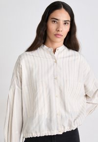 Blusa a righe con bottoni in tessuto leggero. Presenta un colletto alla coreana, due tasche frontali e un elastico in fondo per un fit rilassato.