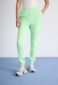 Person som bär ljusgröna joggers, vit linne och vita och grå Nike-sneakers som står på en blå golv.