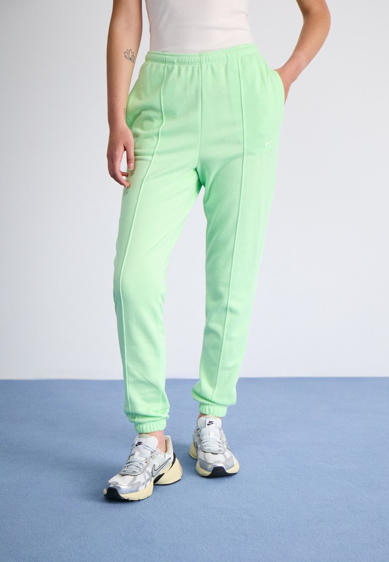 Person som bär ljusgröna joggers, vit linne och vita och grå Nike-sneakers som står på en blå golv.