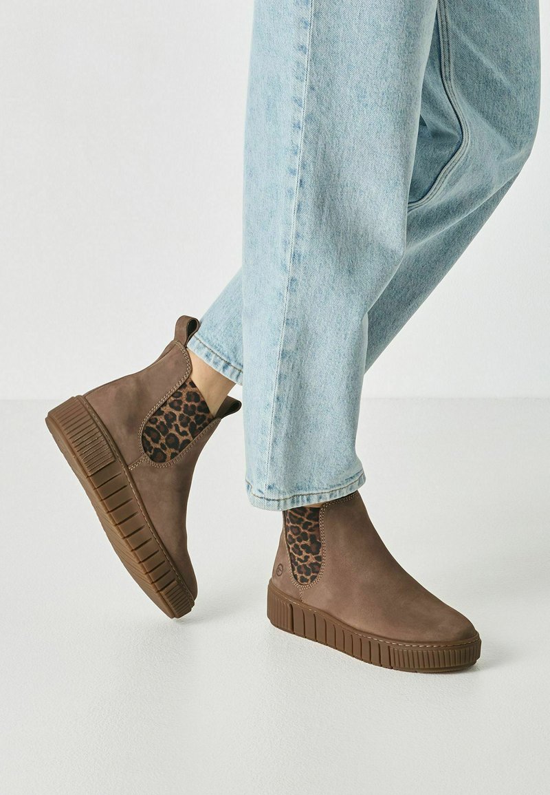 Bottines marron avec panneaux latéraux élastiques à imprimé léopard portées avec un jean bleu clair sur un sol blanc.