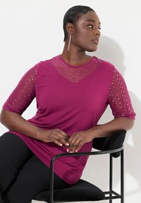 Vibrant lilla top med en blød tekstur, der har gennemsigtige, priklede meshærmer og en V-hals-design. Model sidder på en sort stol.