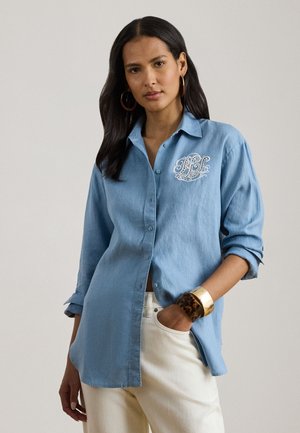 Tummatukkainen nainen pukeutuneena vaaleansiniseen nappipaitaan, jossa on valkoista kirjailua, valkoisiin housuihin, renkaskorvakoruihin ja paksuihin rannekoruihin, poseeraa neutraalisti.