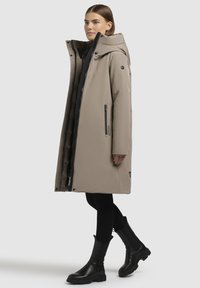 Beige lange jas met een capuchon, zwarte rits en zijzakken; gecombineerd met zwarte enkellaarsjes en zwarte leggings. Gladde, waterafstotende stof.