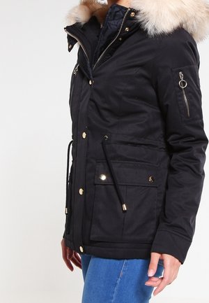 Sort vinterjakke med pelsforet hætte, lynlås foran og trykknapper, lynlåslomme på ærmet og store frontlommer med klap på person iført blå jeans.