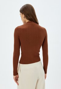 Maglione lungo a maniche lunghe marrone a coste con un design aderente e colletto alto. Tessuto liscio con una stoffa leggermente elasticizzata, abbinato a pantaloni crema.