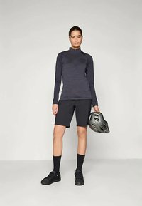 Schwarzes Langarmoberteil mit einem strukturierten Design, kombiniert mit schwarzen Shorts. Die Person hält einen grauen Helm und trägt schwarze Sneakers und Socken.