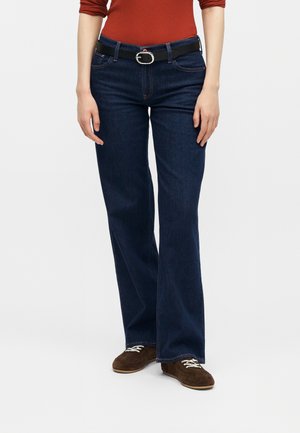 Γυναίκα με σκούρα μπλε καμπάνα τζιν, καφέ παπούτσια από σουέτ, μαύρη ζώνη και μακρυμάνικη μπλούζα σε σκουριασμένο χρώμα, στέκεται μπροστά σε λευκό φόντο.