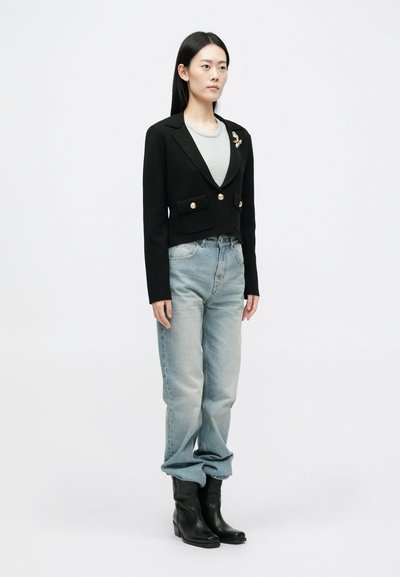 Μαύρο κοντό σακάκι με χρυσά κουμπιά και δύο μπροστινές τσέπες, συνδυασμένο με ανοιχτό μπλε φαρδιά τζιν και μαύρες ankle μπότες. Γκρι πουκάμισο από κάτω.