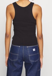 Persona che indossa una canotta nera a costine e jeans blu Lee con tasche applicate, mostrata di spalle su uno sfondo bianco neutro.