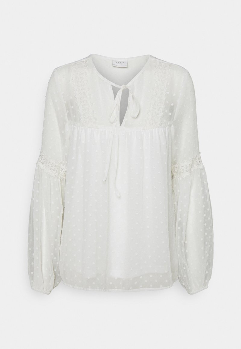 Blouse blanche à manches longues en tissu transparent à pois, détails en dentelle sur la poitrine et les manches, taille froncée, fermeture par lien à l'encolure en forme de goutte.